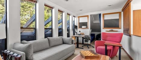 A modern interior with a grey sofa, a red chair, a wooden coffee table with a chessboard, and large windows with a view of trees.