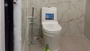 A bathroom with a white toilet, a shower, and a green bucket. - Virat Home Stay (Nivari)