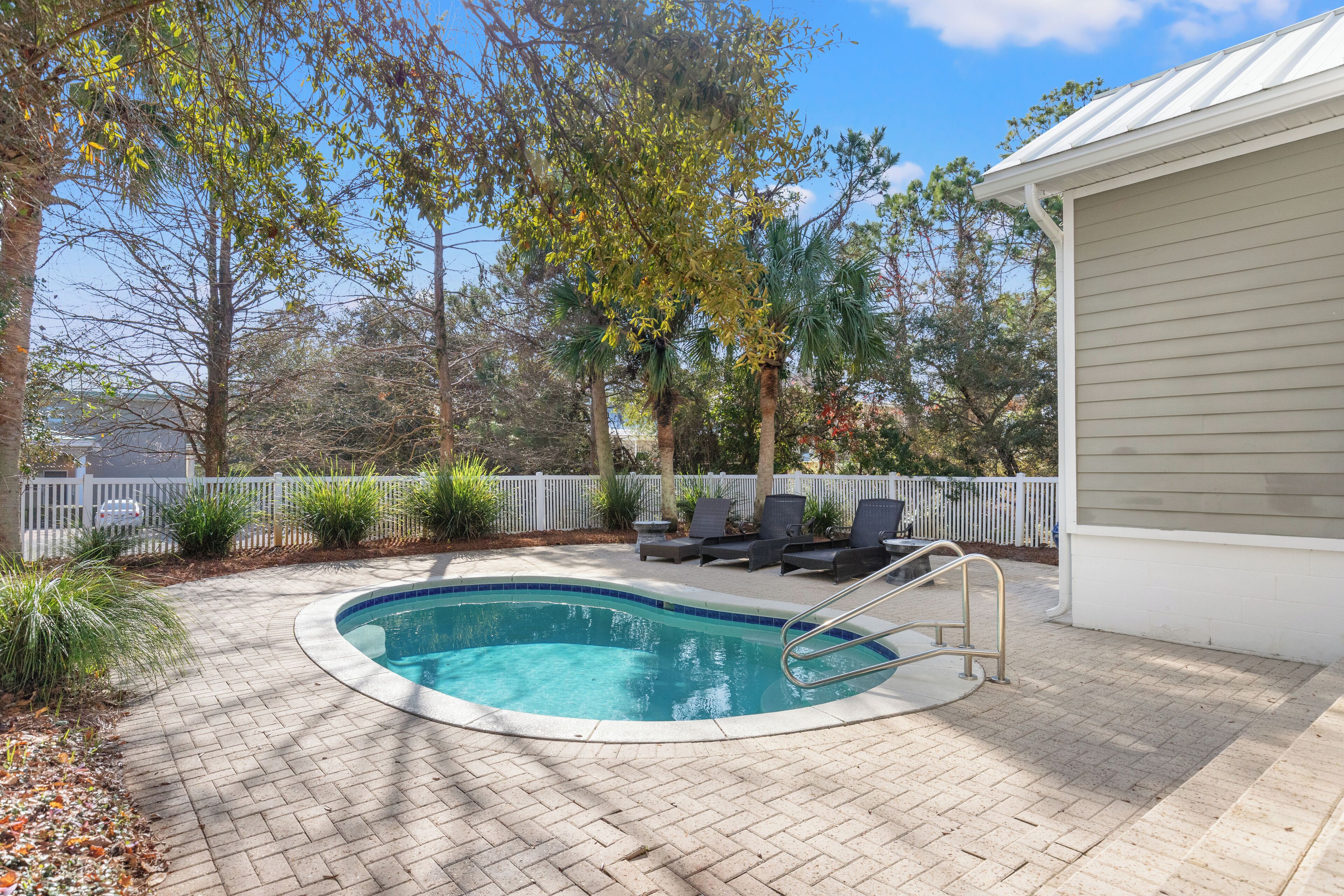 A swimming pool with a brick patio, surrounded by a white fence and various plants.