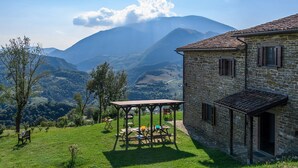 A stone building with a tiled roof, a wooden pergola with picnic tables, and a scenic view of mountains. - Ca Quattrocchi 10&3 by Marche Holiday Villas (Apecchio)