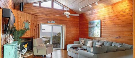 A cozy cabin interior with a wooden floor, a sofa, a chair, a fireplace, and a large window overlooking a body of water.