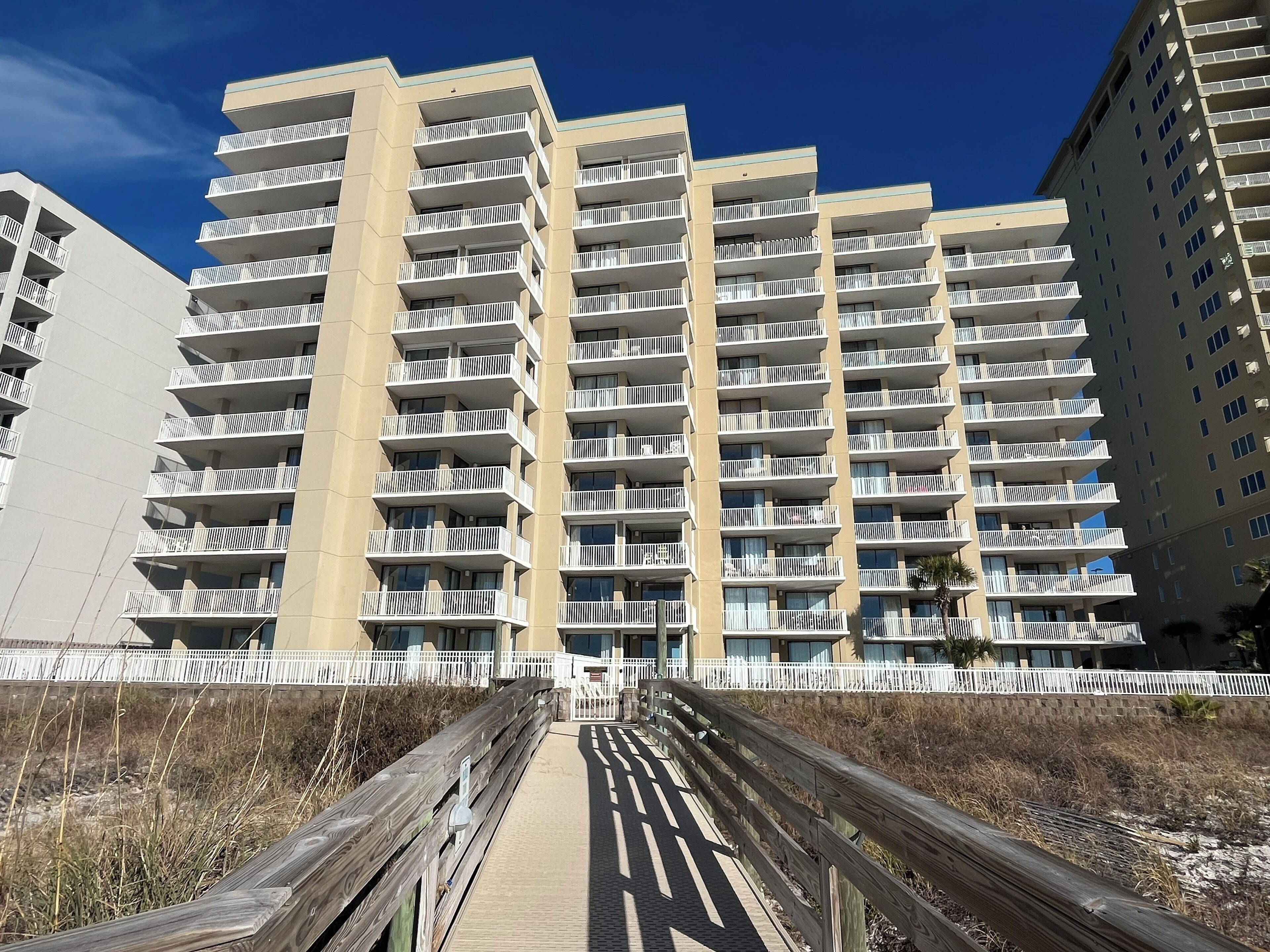 A multi-story residential building with balconies, a wooden boardwalk leading to the entrance, and a clear blue sky.