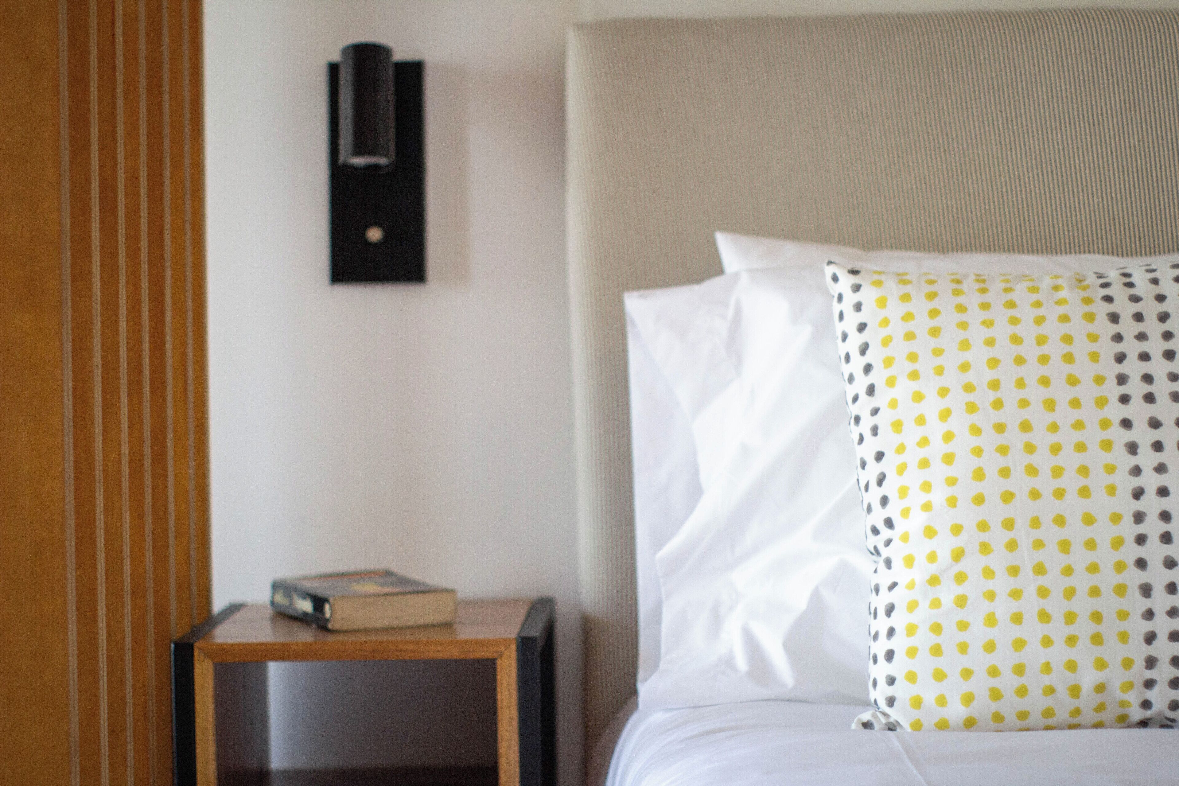 A neatly made bed with white and patterned pillows, a bedside table with a book, and a wall-mounted light fixture.
