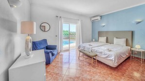 Une chambre d’hôtel avec deux lits, un canapé, une petite table, un ventilateur et une vue sur la piscine, accessible par une porte coulissante.