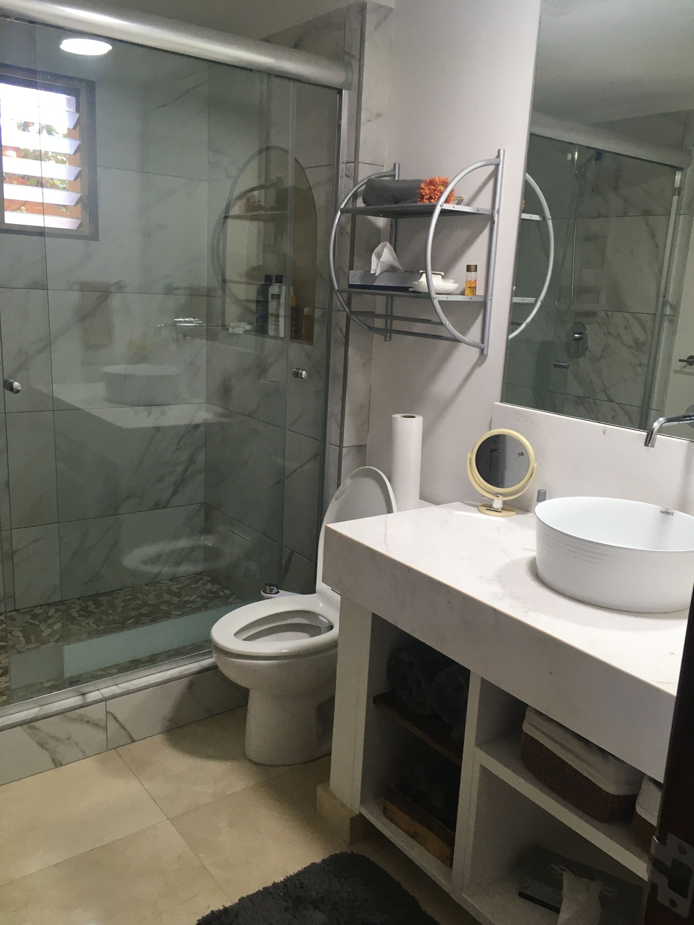 Une salle de bain moderne avec une douche à parois de verre, des toilettes, un lavabo avec un miroir et une étagère contenant des articles de toilette.
