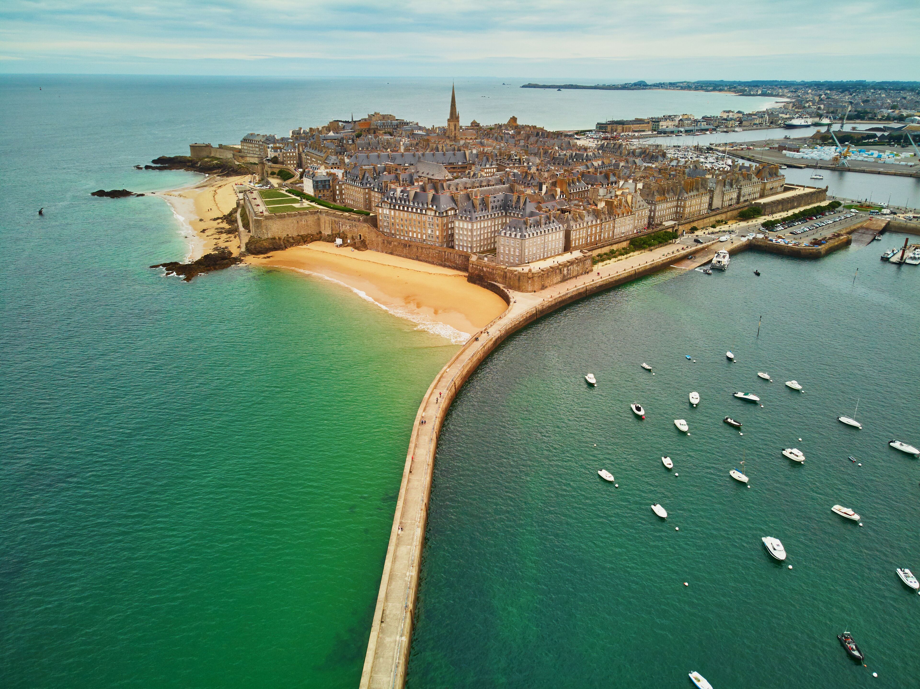 An aerial view of a coastal city with a prominent castle, a sandy beach, and a marina with numerous boats.