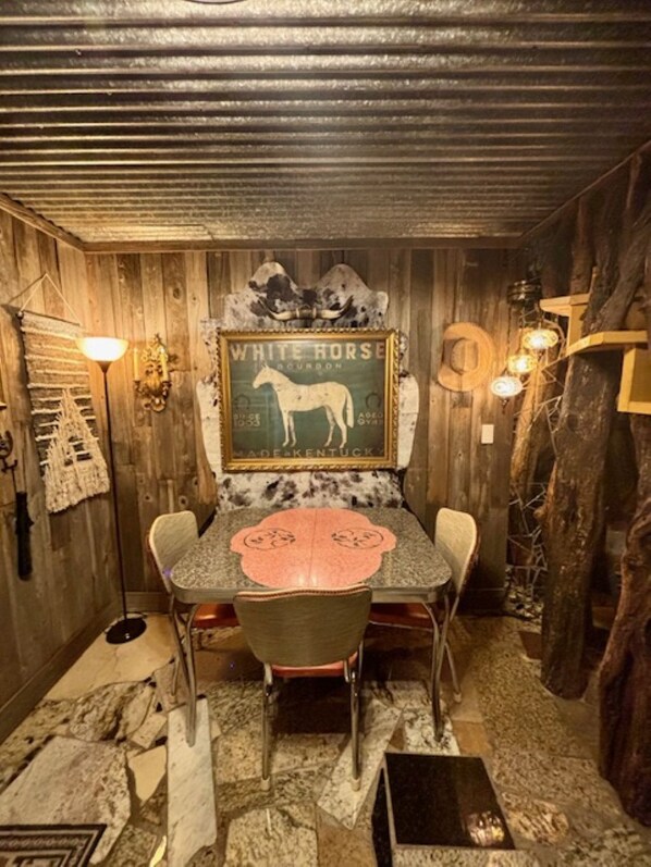 A rustic room with a wooden ceiling, a table with chairs, a framed picture of a white horse, and a wall-mounted light fixture. - The World-Famous Cowboy Hat House (Huntsville)