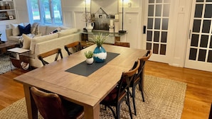 A dining area with a wooden table, chairs, and a vase with plants. There is a glass-top coffee table, a sofa, and a dining table with chairs. The room has a door with frosted glass panels and a window with a view of trees. - A Step Away: The Retreat – Walk to Town Escape (Saugatuck)