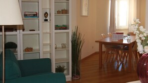 A living room with a teal sofa, a dining table with chairs, a bookshelf, and a window with curtains.