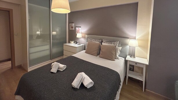 A hotel room with a large bed, two bedside tables with lamps, a glass door, and a grey carpet.