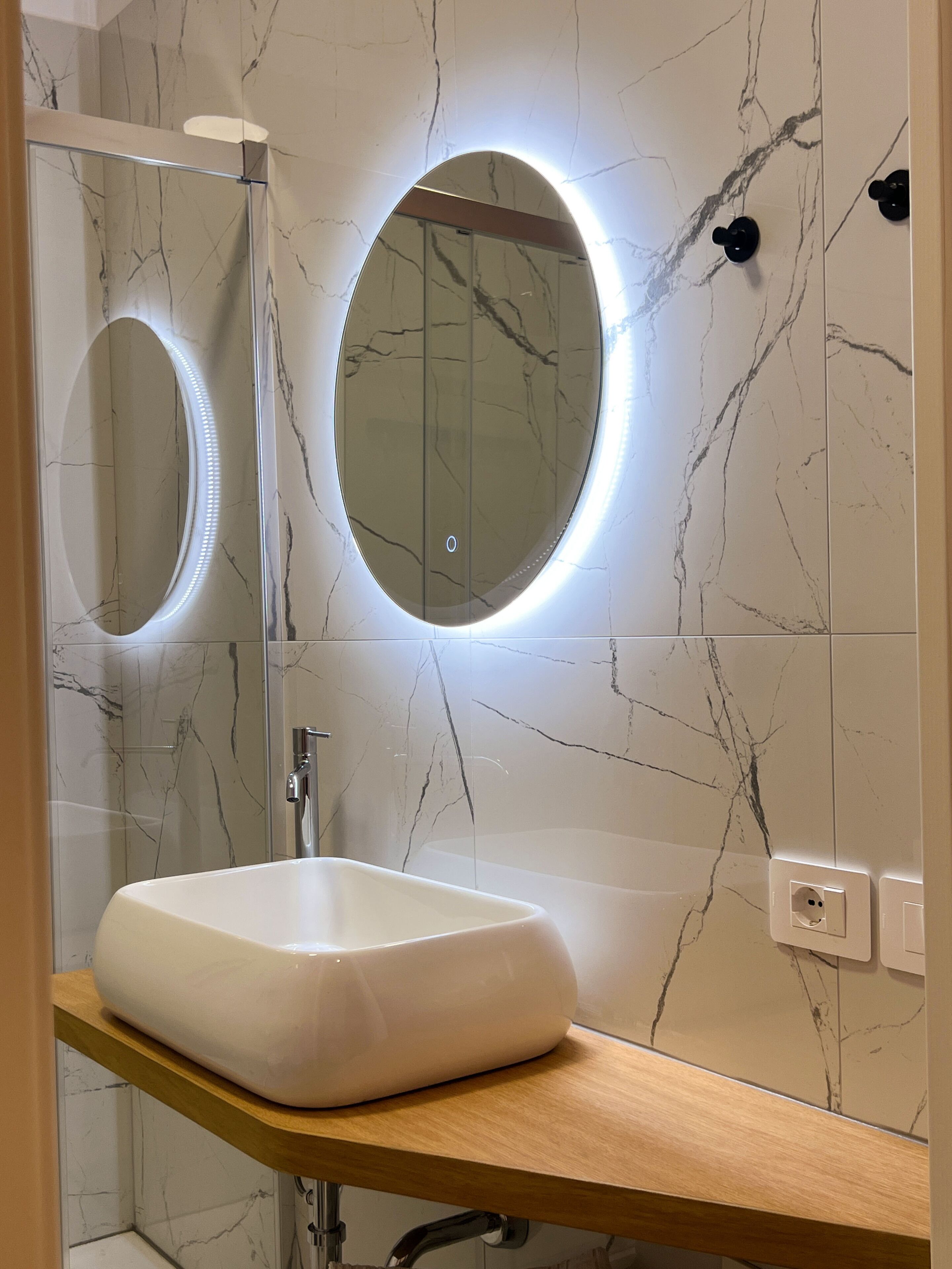 A bathroom with a white marble sink, a mirror with a circular light, and a shower area.