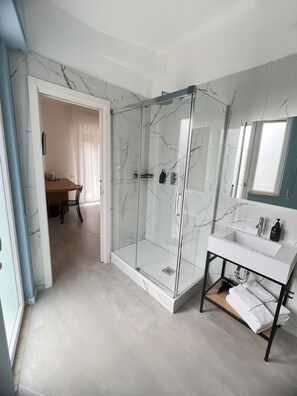 A modern bathroom with a glass-enclosed shower, a white sink with a marble countertop, and a wooden bench. - Le camere del centro (Matera)