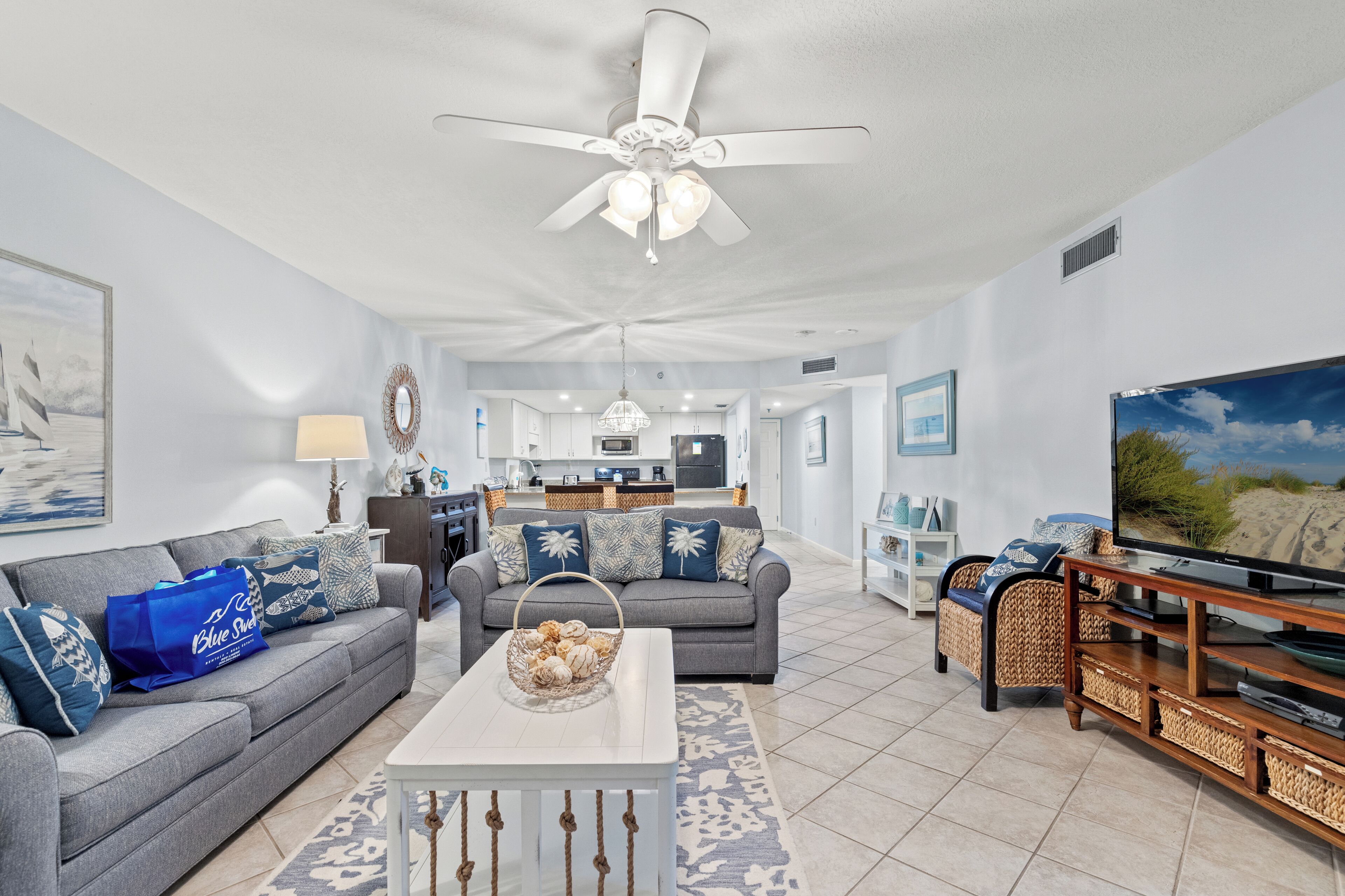 A spacious living room with a grey sofa, a white coffee table, and a television. There is a ceiling fan, a kitchen in the background, and a dining area.