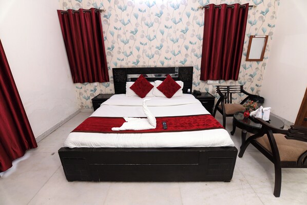A neatly made bed with a white and red bedspread, two red pillows, and a white swan decoration. There is a small table with a chair beside the bed, and a wall-mounted mirror. - Hotel Ashray Noida (Noida)