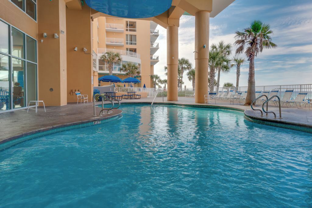 An indoor swimming pool with lounge chairs and umbrellas, surrounded by palm trees and a multi-story building with balconies.