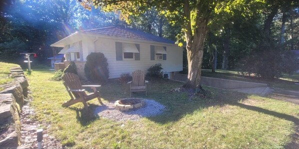 A single-story house with a front porch, a fire pit, and a tree in the yard.