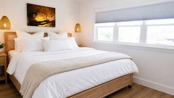 A bedroom with a wooden bed, white bedding, a painting on the wall, and a window with blinds.