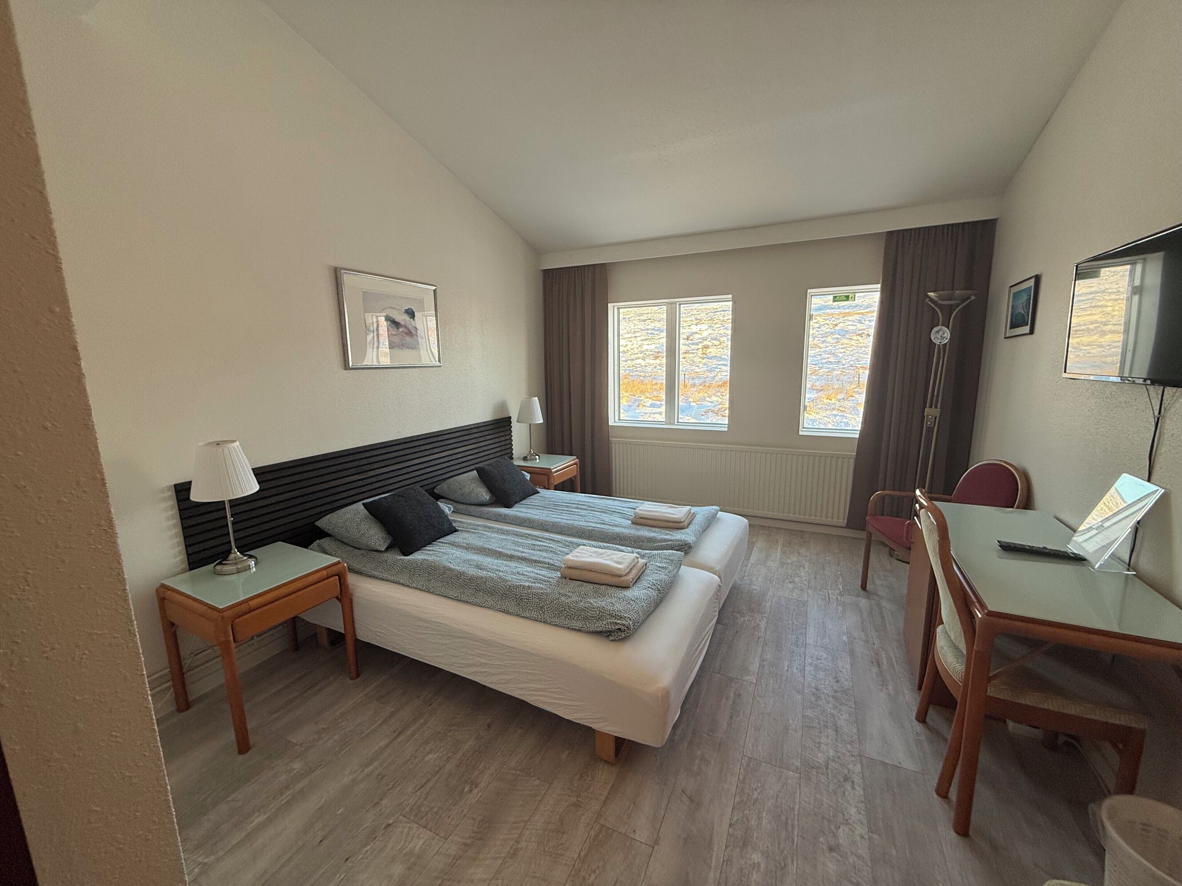 Camera d'albergo con due letti, una scrivania, una sedia, televisione e finestre con tende.