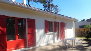 Una casa con contraventanas rojas, fachada blanca y una terraza de madera con mobiliario exterior.