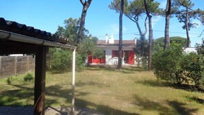 Una casa con puerta roja, pared blanca y chimenea. Hay árboles y un patio con césped al frente.