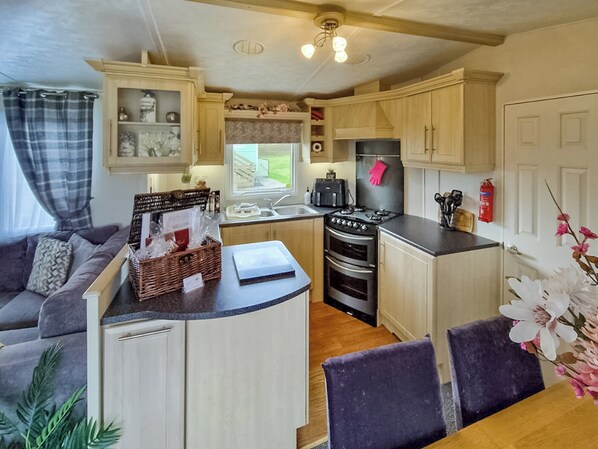 A compact kitchen with a central island, a dining area with a table and chairs, and a living area with a sofa and a plant. - Roe Meadow 3 - UK48411 (Newton Stewart)