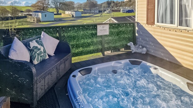 Un jacuzzi con panel de control, un sofá de mimbre con cojines y vistas a un paisaje verde con edificios al fondo.