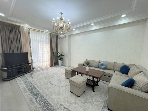 A living room with a sofa, ottomans, a coffee table, a TV, and a chandelier. - New Kamolon by RNB estate (Tashkent)