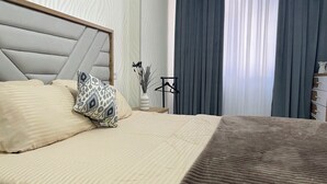 Ein ordentlich gemachtes Bett mit Kopfteil, zwei Kissen, einem Nachttisch mit einer Vase und einem schwarzen Kleiderbügel sowie einem Fenster mit Vorhängen.