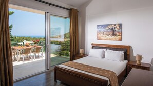 A hotel room with a large bed, a painting on the wall, a balcony with a table and chairs, and a view of the sea. - Villa Vlychada w. Pool, Jacuzzi & Sea Views (9QXM+554 Vlichada, Greece)