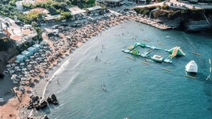A coastal town with buildings on a hillside, a beach with umbrellas, and boats in the water. - Villa Vlychada w. Pool, Jacuzzi & Sea Views (9QXM+554 Vlichada, Greece)
