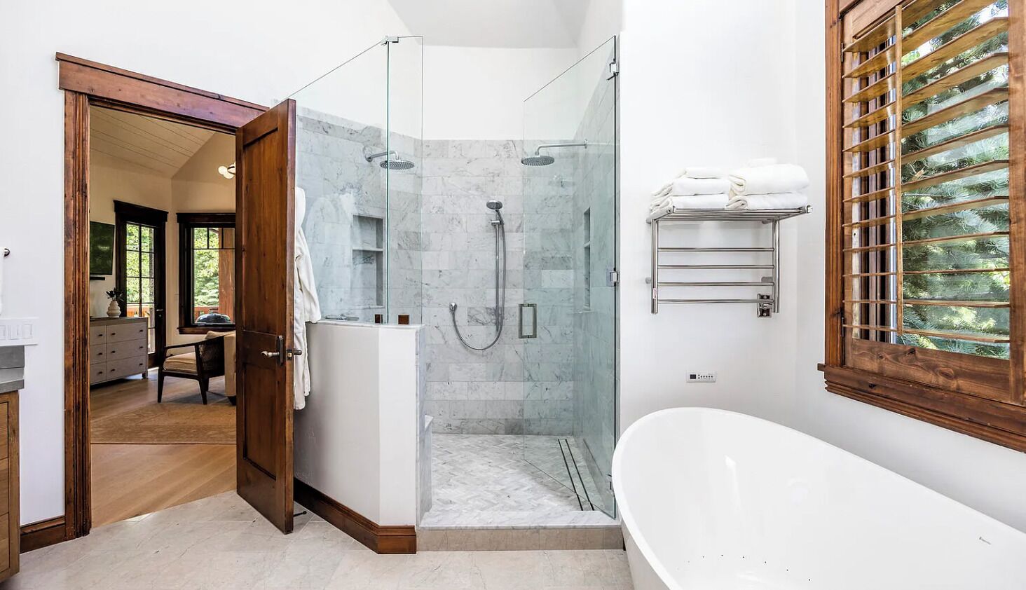 A modern bathroom with a glass-enclosed shower, a freestanding bathtub, and a wooden window with blinds.