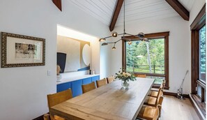 Un espace repas avec une longue table en bois, des meubles de rangement bleus et une grande fenêtre donnant sur un paysage verdoyant.
