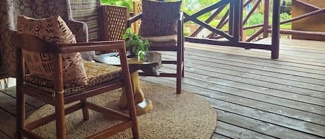 A wooden deck with thatched roof, furnished with chairs and a table, overlooking a tropical landscape.