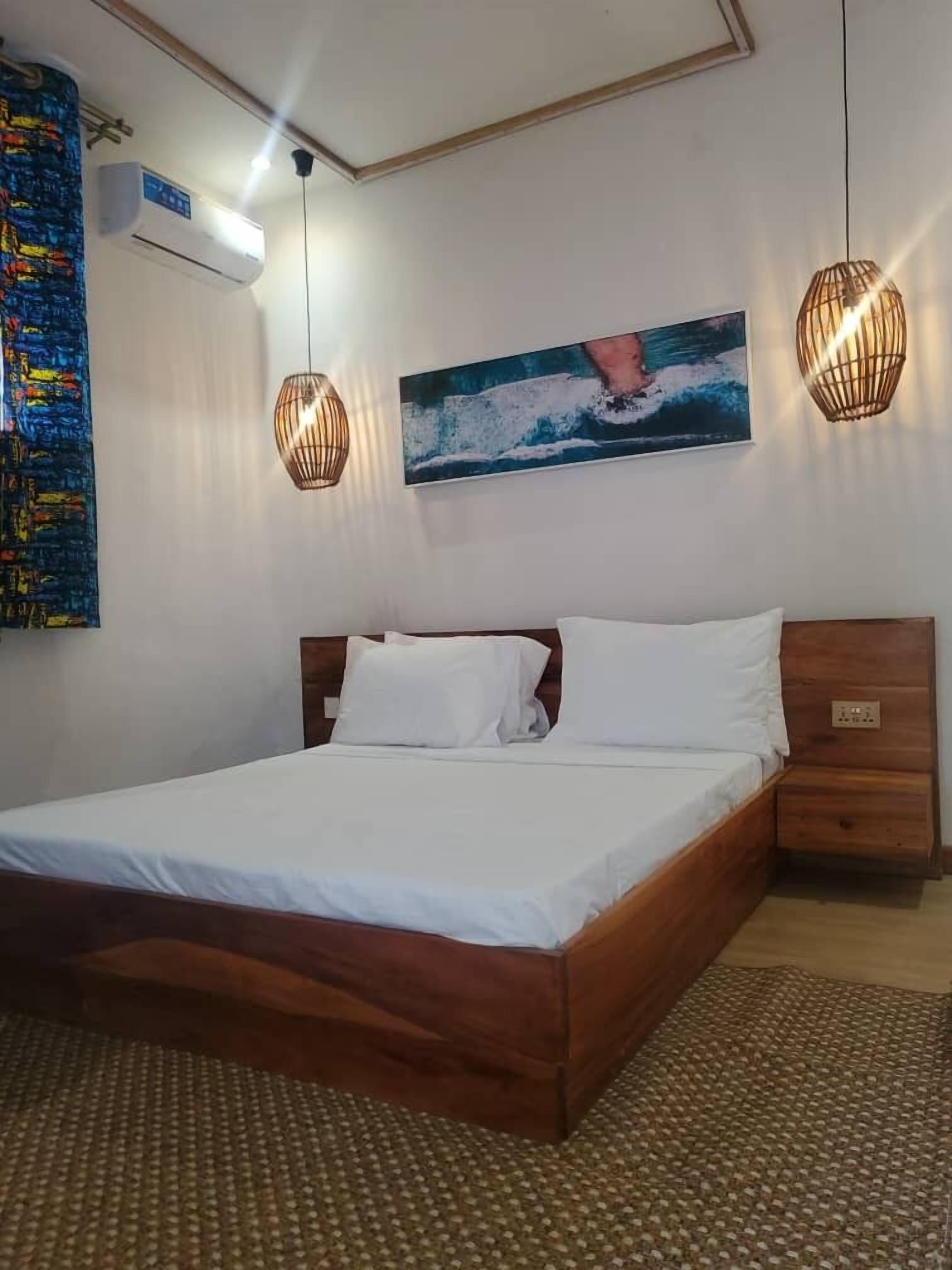 Un dormitorio con cama de madera, ropa de cama blanca, un cuadro de una persona surfeando, dos lámparas colgantes y un aire acondicionado instalado en la pared.