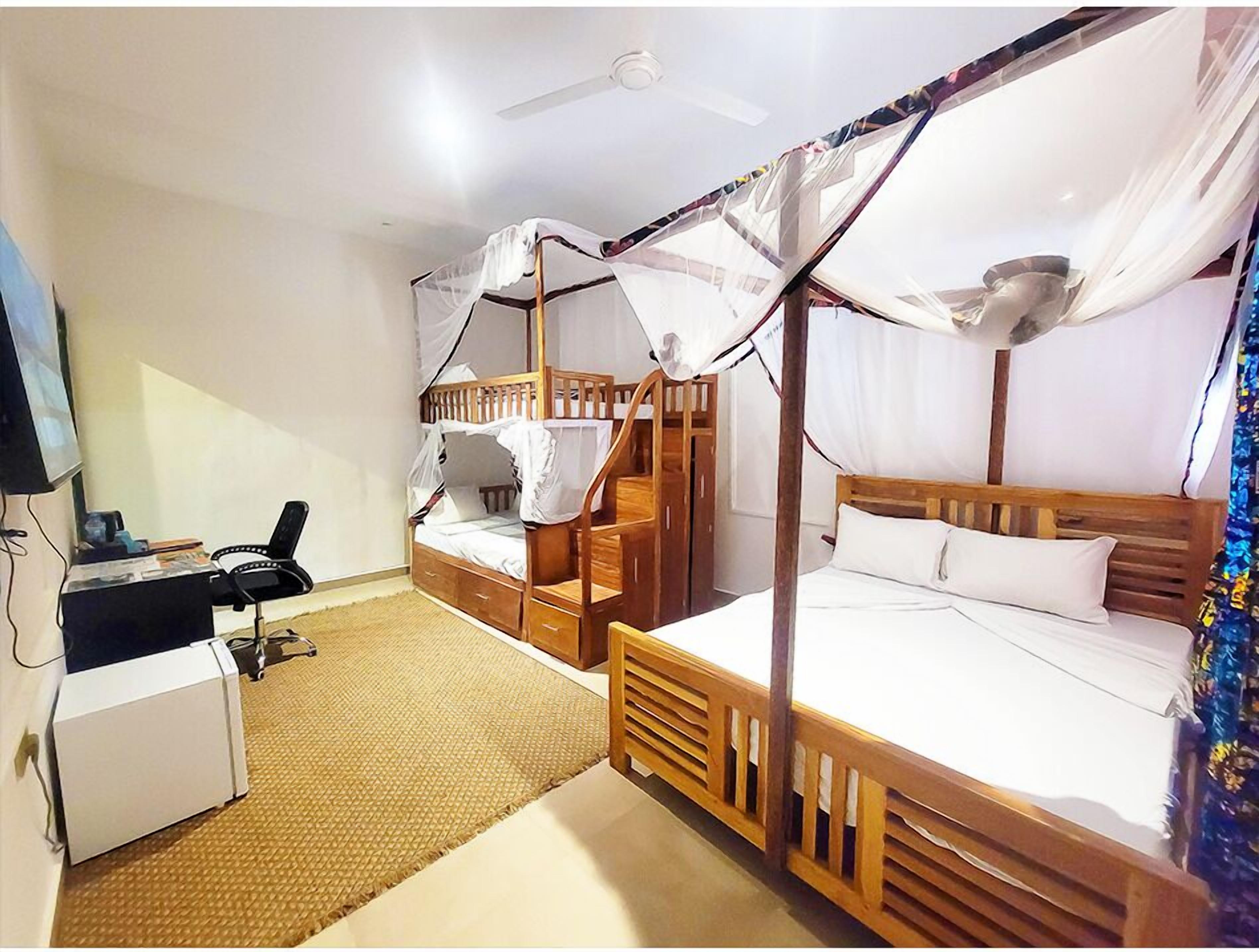 Una habitación con dos literas de madera, un escritorio con silla, un armario blanco pequeño y un ventilador de techo.