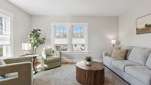 Una sala de estar con un sofá gris, una mesa de centro redonda de madera, un sillón verde y una lámpara blanca. Hay dos ventanas con persianas, una planta y un cuadro en la pared.