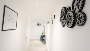 A hallway with a framed artwork on the left, a clock on the wall, and a chair with a blue cushion. - Stylish London Retreat | Spacious & Modern Home for 10 Guests (Greater London)