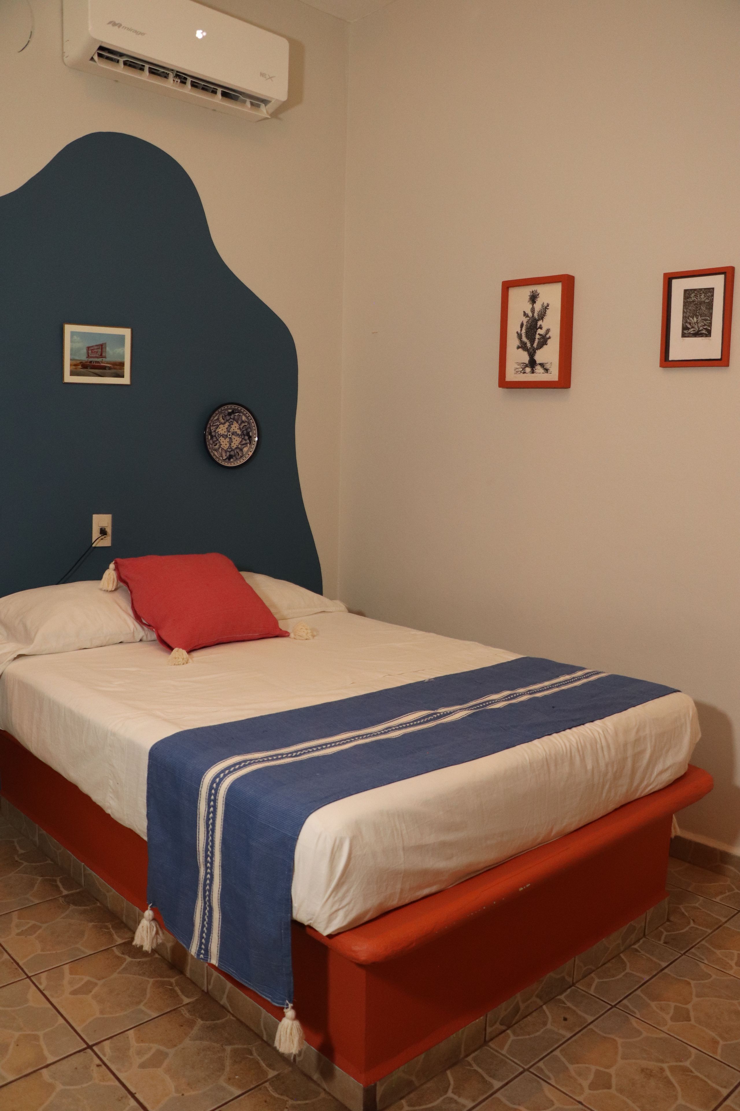 A bedroom with a bed, a red headboard, a wall-mounted air conditioner, and two framed pictures.