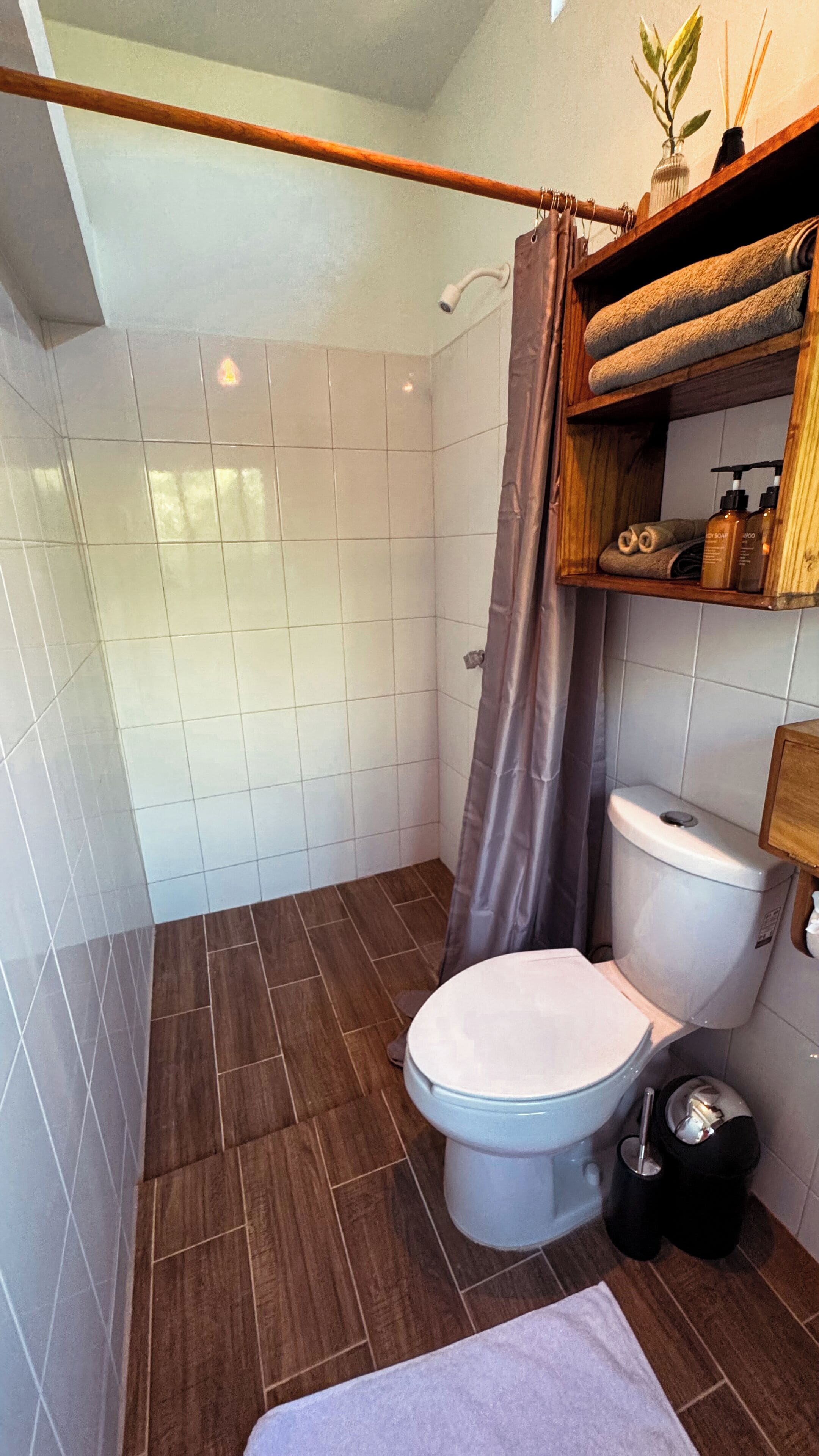 Un baño con inodoro, cortina de ducha, repisas con artículos de aseo y una toalla en el suelo.