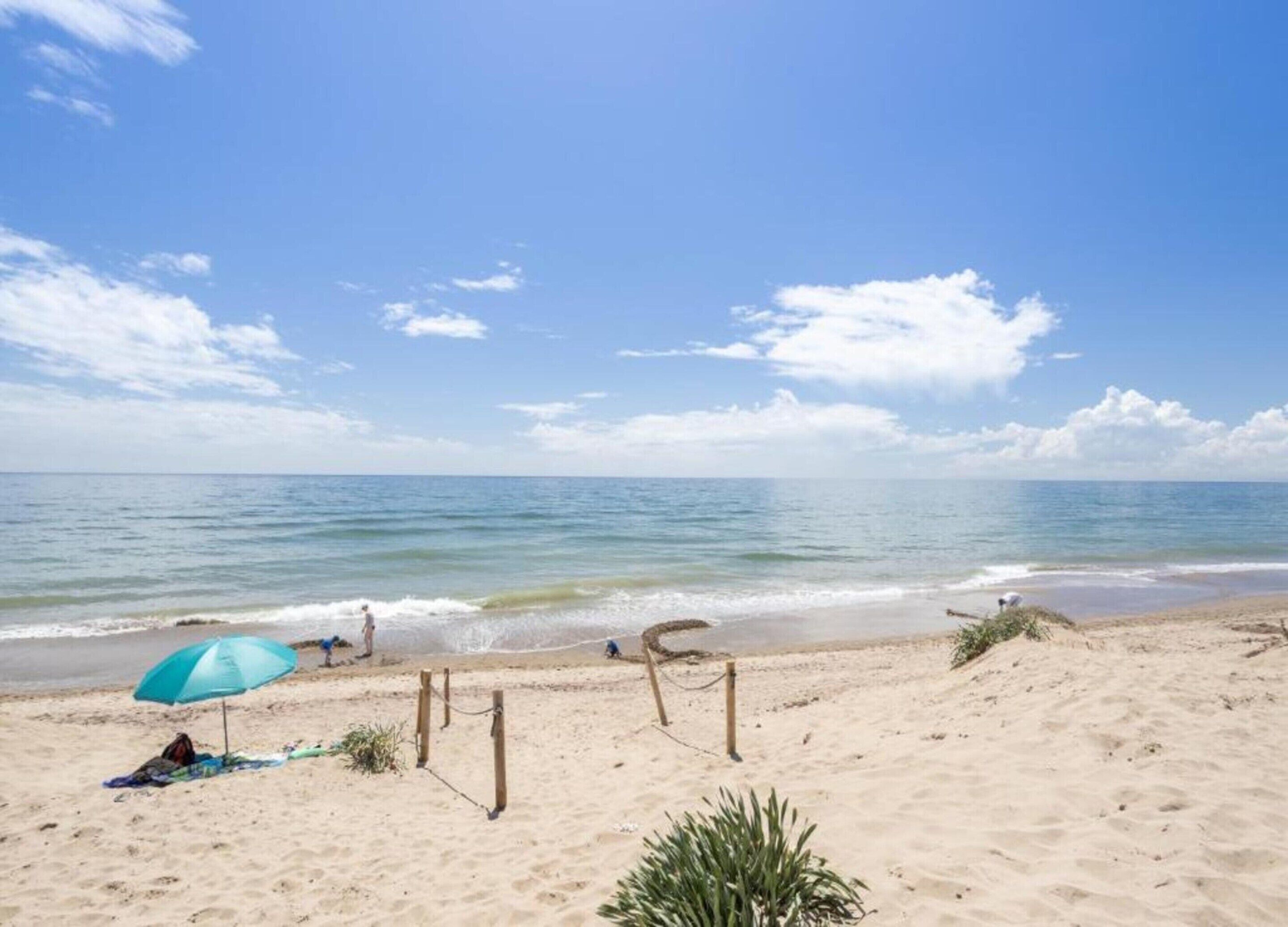 Une plage avec un ciel dégagé, un rivage sablonneux et une personne abritée sous un parasol bleu.