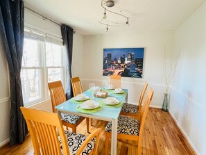 Un coin repas avec une table dressée pour quatre personnes, des chaises en bois ornées de coussins à motifs, un tableau représentant un paysage urbain accroché au mur et un vase avec des branches posé sur le sol.