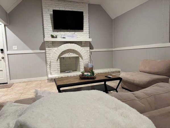 Una sala de estar con chimenea de ladrillo blanco, un televisor de pantalla plana montado encima, un sofá beige y una mesa de centro con una cesta de plantas.