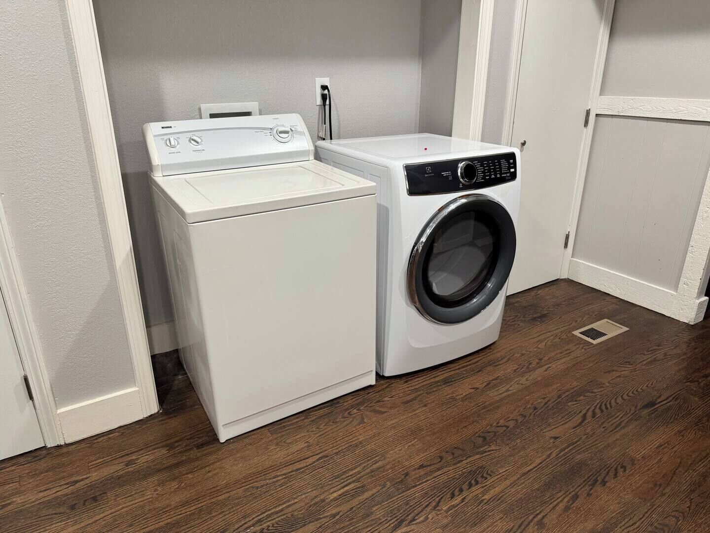 A laundry room with a front-loading washer and dryer, wooden flooring, and white walls.
