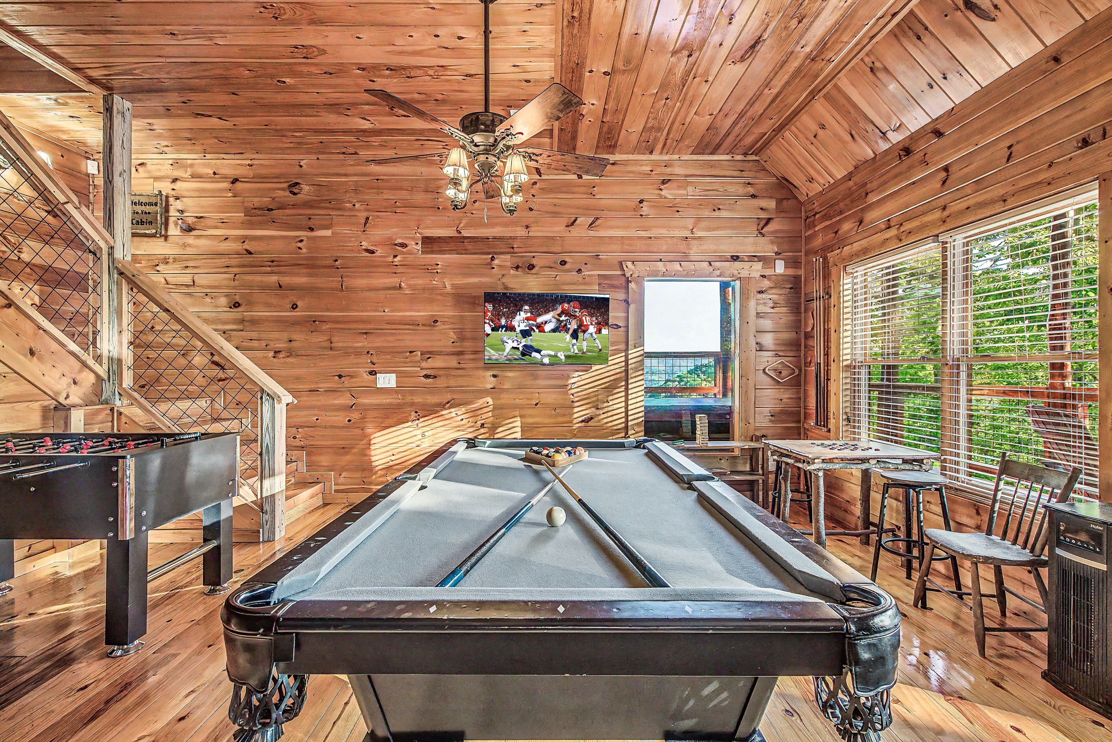 A pool table in a wooden room with a ceiling fan, a foosball table, and a window with blinds.