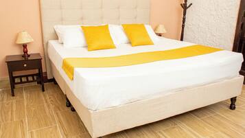 A neatly made bed with white and yellow bedding, a wooden nightstand with a lamp, and a wooden coat rack against a peach-colored wall.