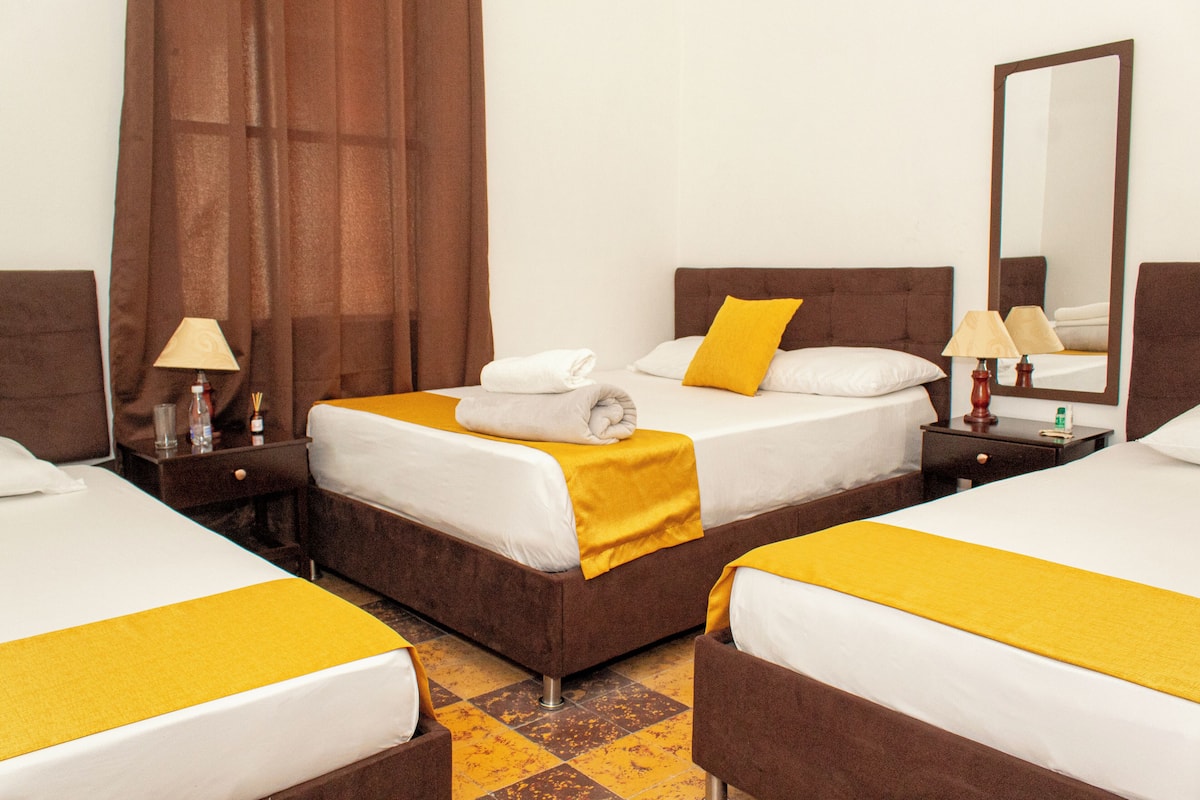 Habitación de hotel con dos camas, cada una con cabecero marrón, ropa de cama blanca y una almohada decorativa amarilla. Hay dos mesitas de noche con lámparas y un espejo en una pared.