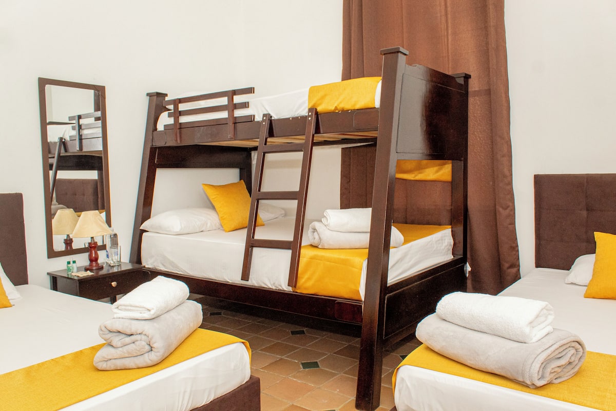 Habitación de hotel con dos camas individuales, una litera, un espejo, una mesita de noche con lámpara y un baño.