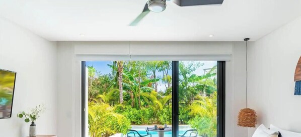 A modern room with a large sliding glass door leading to a pool area surrounded by lush greenery.