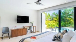 A modern hotel room with a bed, a desk, a chair, a TV, and a view of a pool and greenery. - Super Studio Laguna C 106 (Tulum)