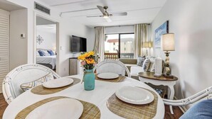 Un coin repas avec une table ronde dressée pour quatre personnes, des chaises blanches, un vase avec des fleurs et un ventilateur de plafond.